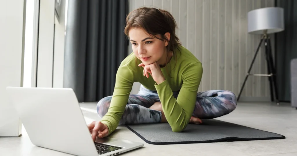 Femme concentrée regardant un ordinateur en position assise sur un tapis de yoga, illustrant l’apprentissage du yoga en ligne.